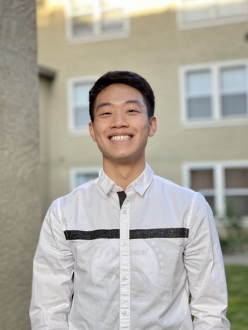 A young asian man with short dark brown hair wears a white long-sleeved button down with a black line across the chest and smiles in front of a light yellow building.